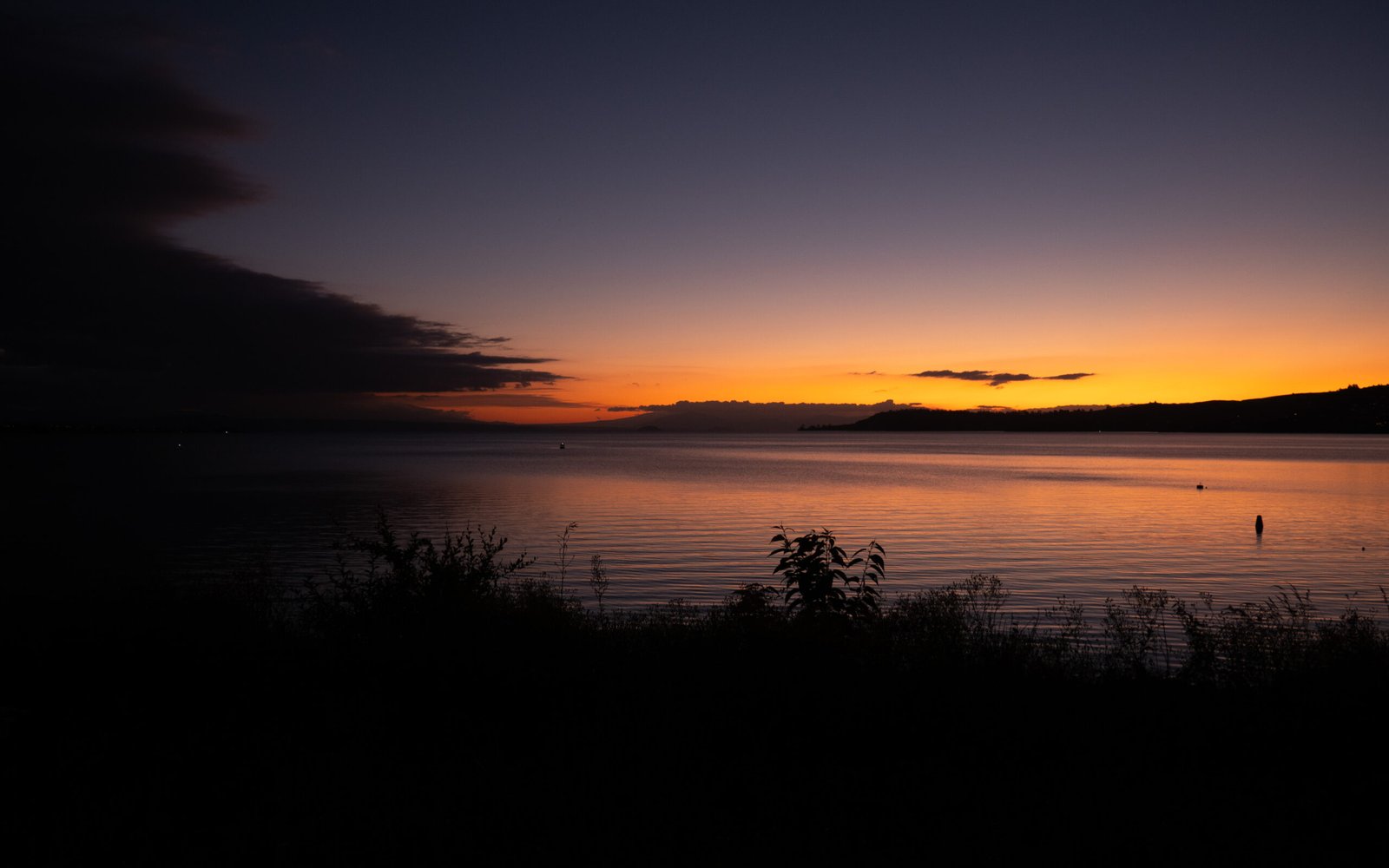 Photography - A serene sunset over a calm body of water is accentuated by the glowing hues of the twilight sky, creating a tranquil and picturesque scene. Silhouettes of distant hills and foliage in the foreground add to the sense of peacefulness. Lake Taupo, NZ.