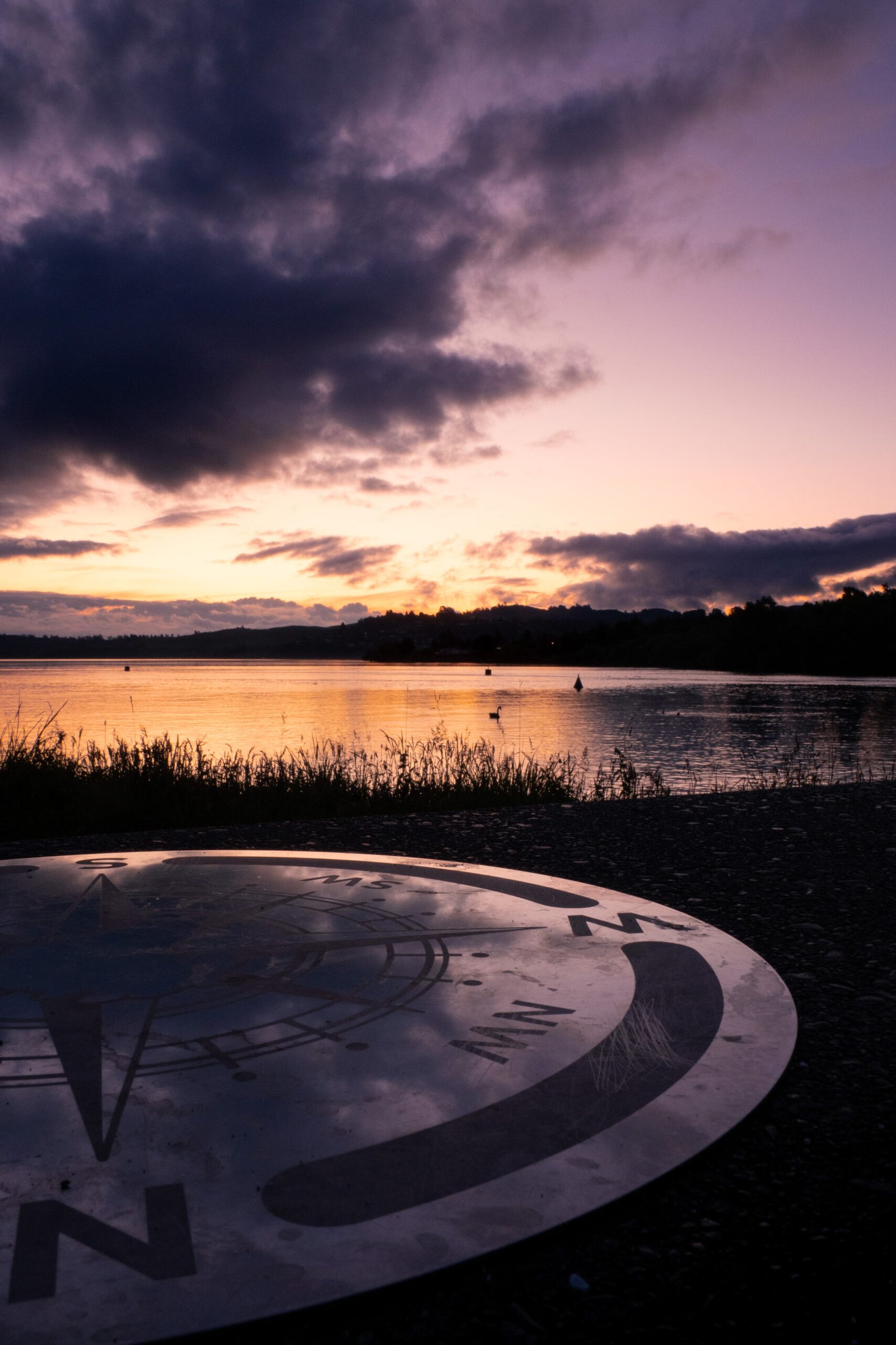 Photography - A compass rose is laid out on the ground with a serene lake and vibrant sunset in the background. Dark clouds partially cover the sky, adding dramatic contrast to the colourful horizon. Lake Taupo, NZ.