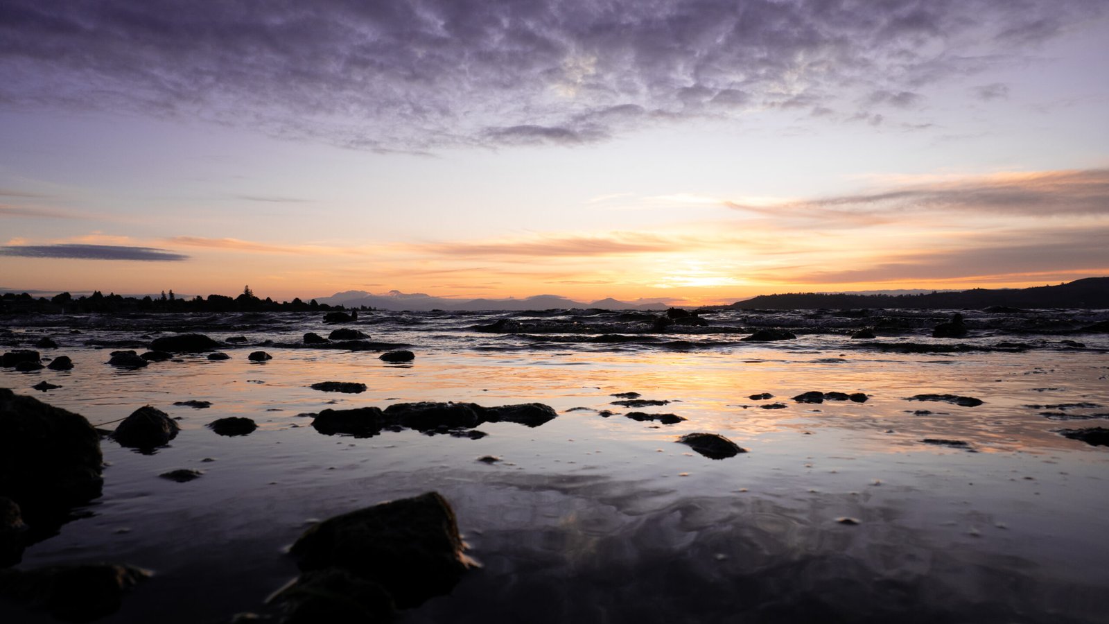 Photography - A tranquil seascape captures the serene beauty of a sunset, with the horizon painted in hues of orange and purple. Silhouetted rocks and gentle waves add depth to the reflective water's surface.